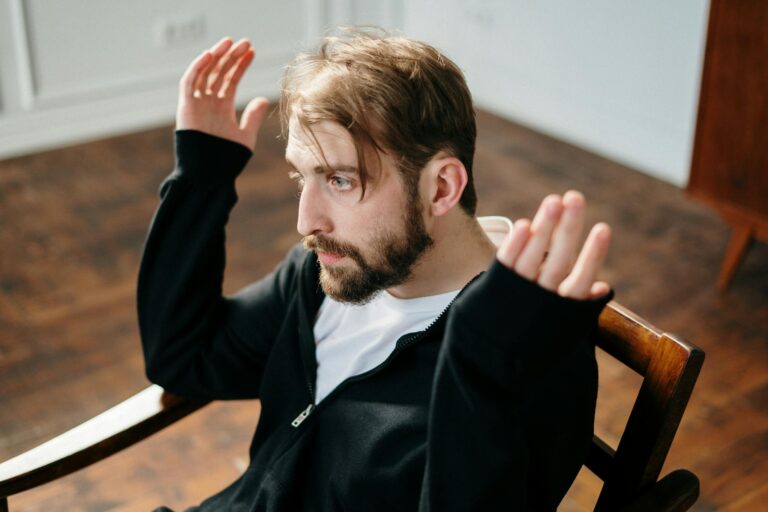 Bearded man sitting and gesturing expressively in a well-lit interior space.